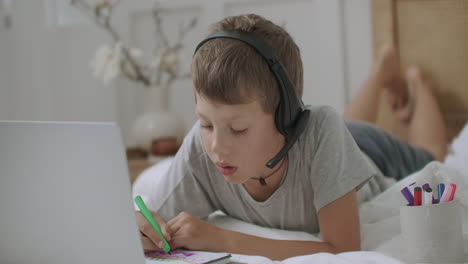 little boy is sitting on couch and drawing on paper using laptop with video tutorial listening by headphones