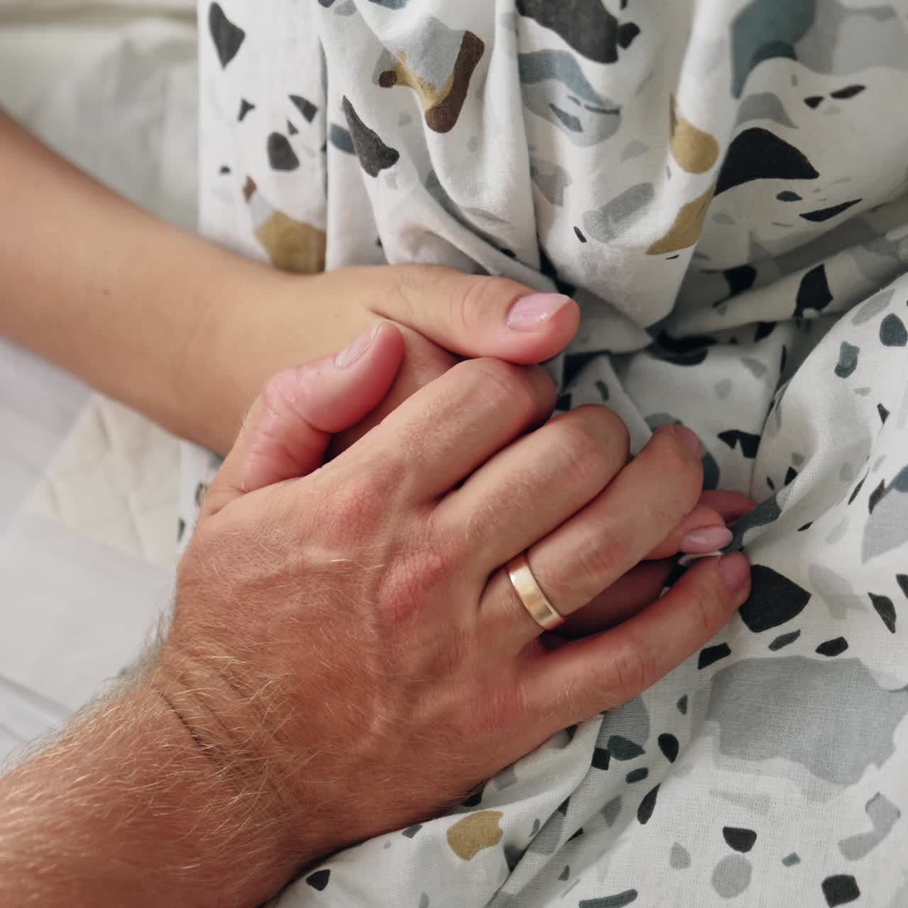 Male hand with a wedding ring covering the female one. Spouses' hands lying on the bedding. Close up