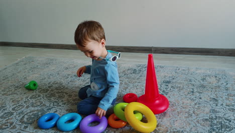 cute kid wearing blue shirt and pants sits on the floor. kid collects pyramid and waves hands energetically.
