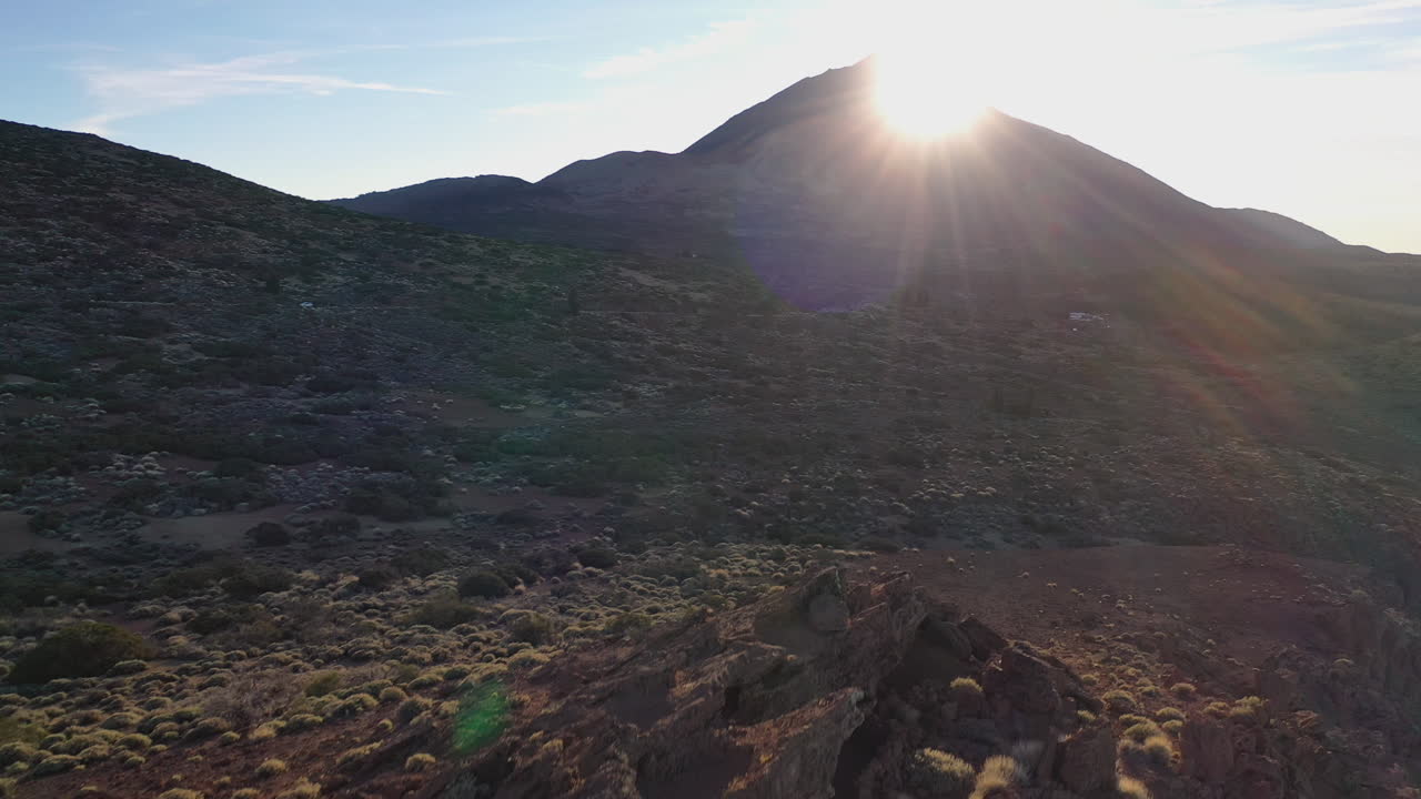 The Sun setting behind the summit of Pico de Teide on Canary Islands with a rocky hillside with bushes below lit by sunrays. The Sun setting behind a large mountain shining on the valley below 4K.