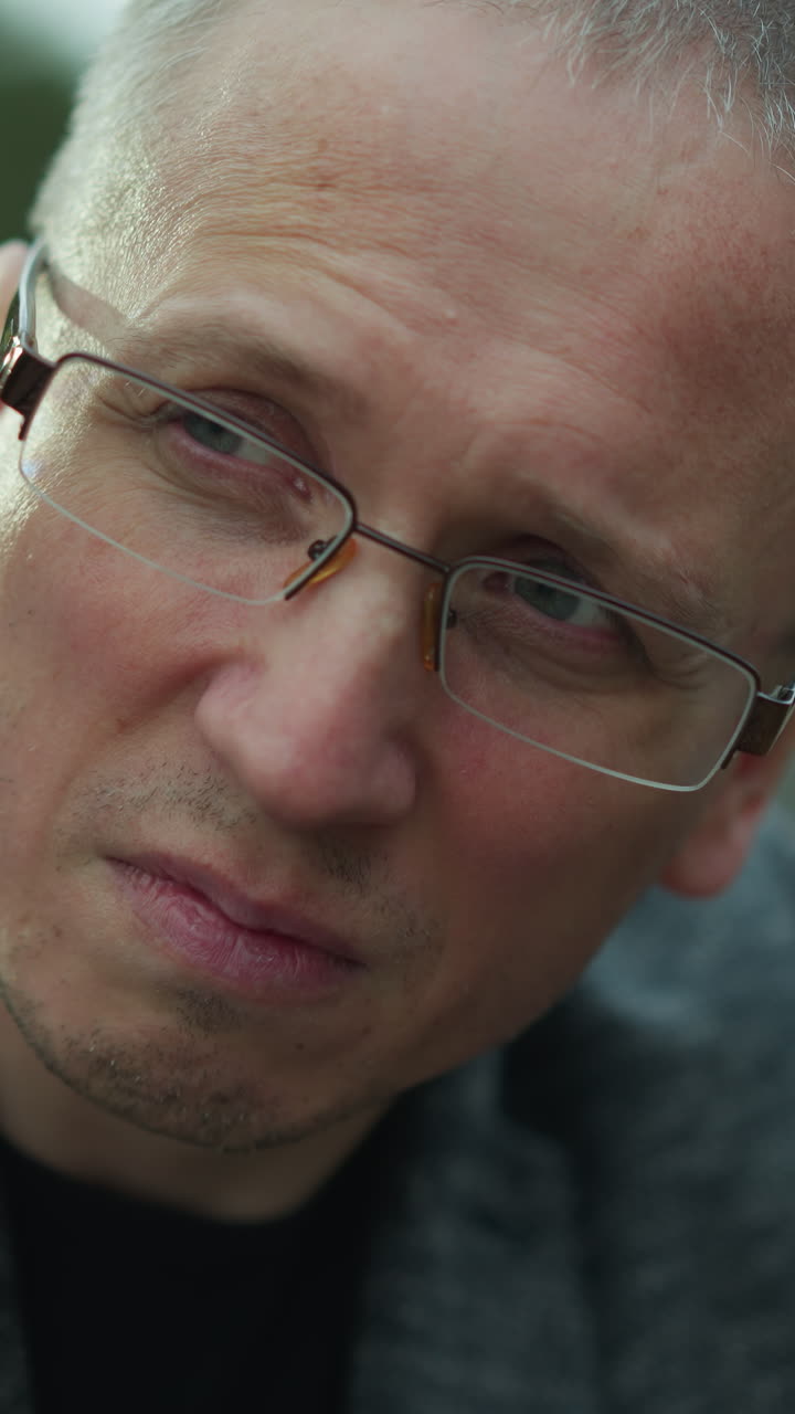 una vista de cerca de un hombre de mediana edad en gafas con una expresión seria, está usando una chaqueta gris y está al aire libre con un fondo borroso de árboles