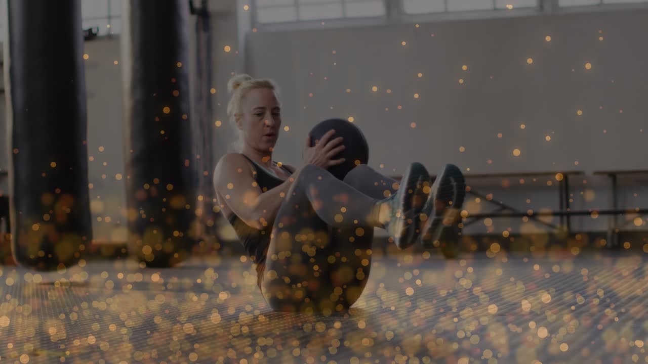 Woman starting V-sit and rotating medicine ball in gym, golden bokeh highlighting motion for core