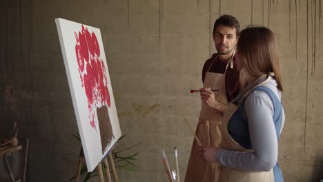 Female-artist-teaching-young-male-student-to-paint-with-oil-paints.-Dark-haired-man-in-beige-apron-drawing-pink-flowers-using-paintbrush.-Workshop-in-art-studio.-Slow-motion