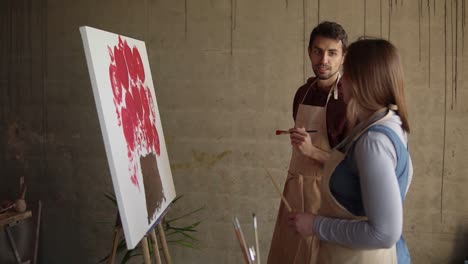 Artista-Femenina-Enseñando-A-Un-Joven-Estudiante-A-Pintar-Con-Pinturas-Al-óleo.-Hombre-De-Pelo-Oscuro-Con-Delantal-Beige-Dibujando-Flores-Rosadas-Con-Pincel.-Taller-En-Estudio-De-Arte.-Camara-Lenta