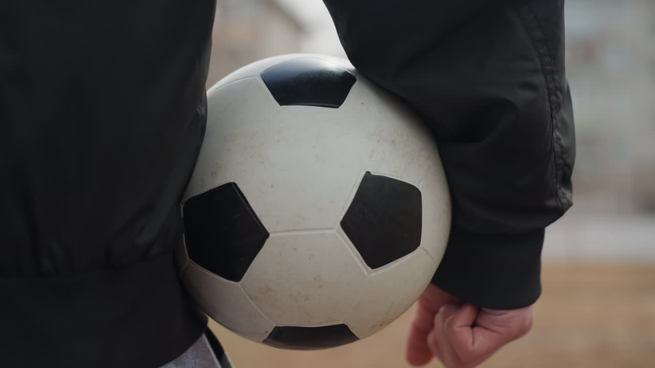 Tæt beskåret billede af en fodbold klemt ind mod hoften. En hvid spiller står ved siden af målet på en lokal bane med en afslappet holdning. Den eftertænksomme stemning antyder en pause i halvlegen eller en plan før genstart