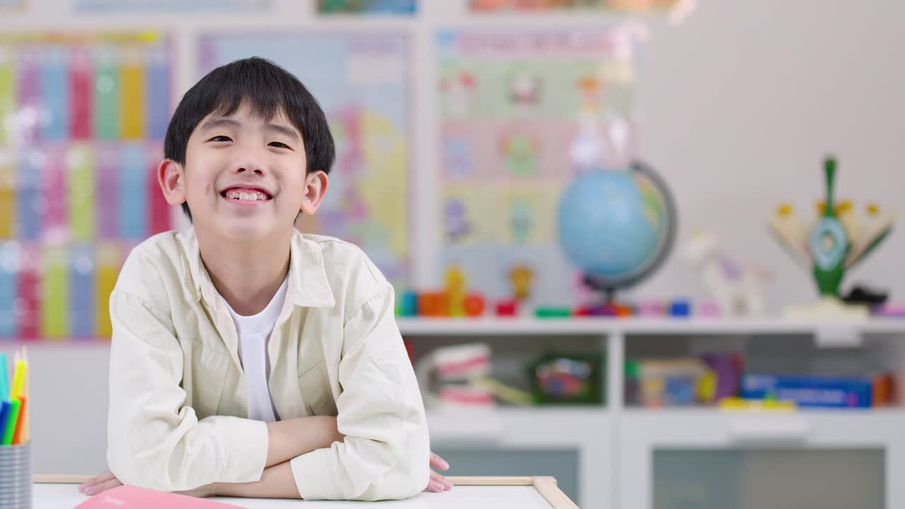 明るくカラフルな教室で、腕を組み机に向かって微笑む幸せそうな男の子