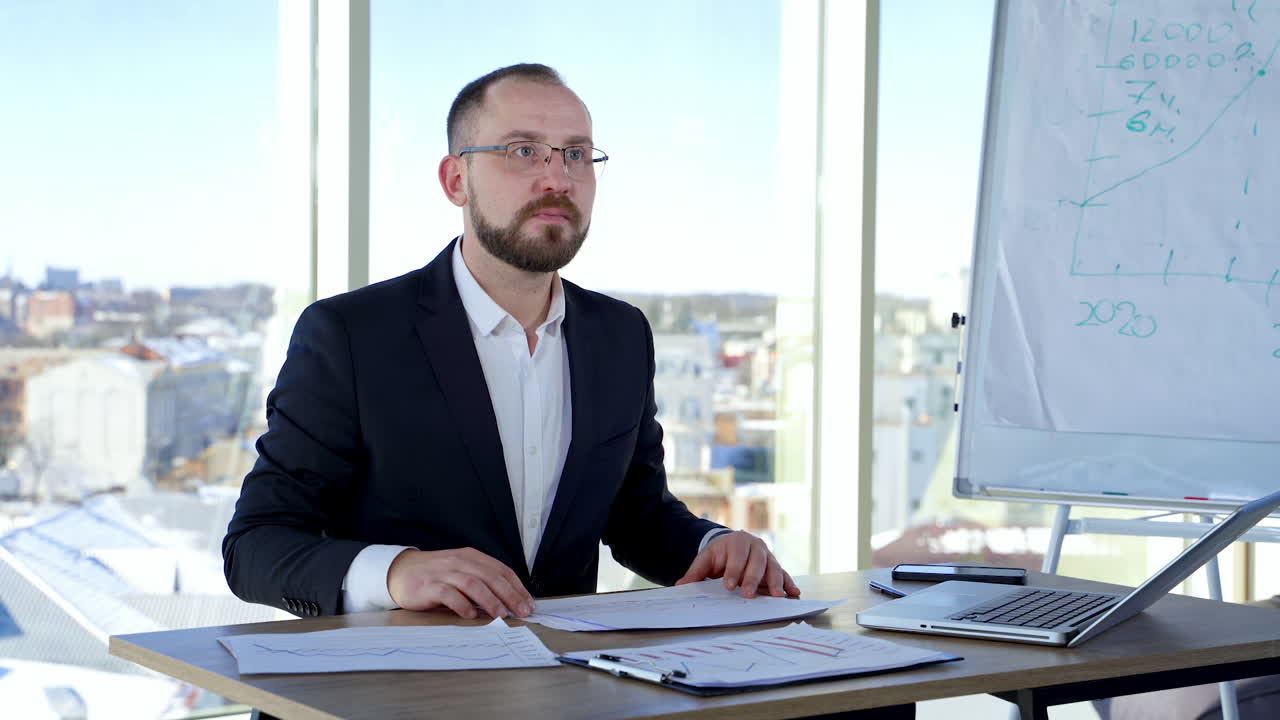 Businessman in glasses at his workplace. Executive man in suit sitting at desk with many papers and work in office against window background.