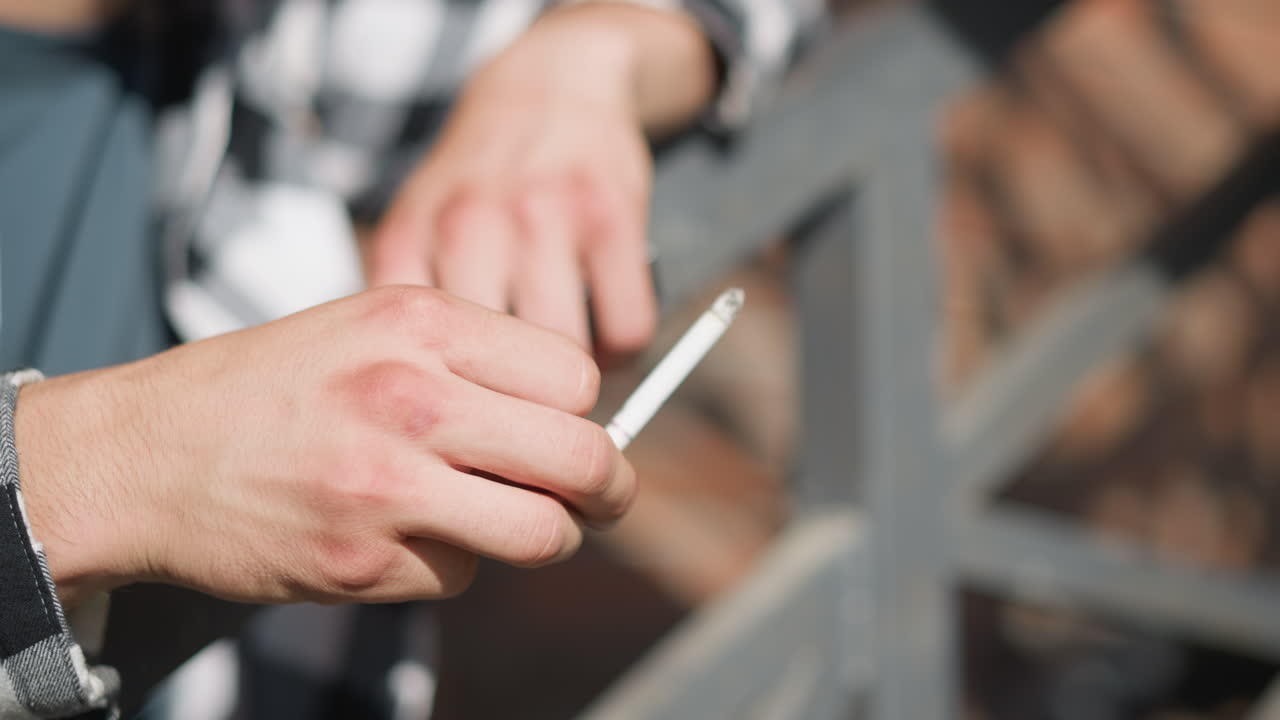 close up of white man hand holding cigarette while smoking near metal railing in sunlight with soft blur background and checkered shirt slightly visible during relaxed moment outdoors