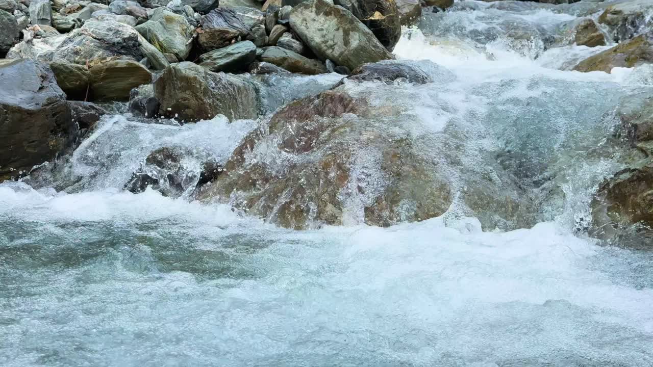 Fast-moving glacial meltwater rushes over rocks in a mountain stream, captured in natural daylight with a steady, fixed camera angle and crisp detail