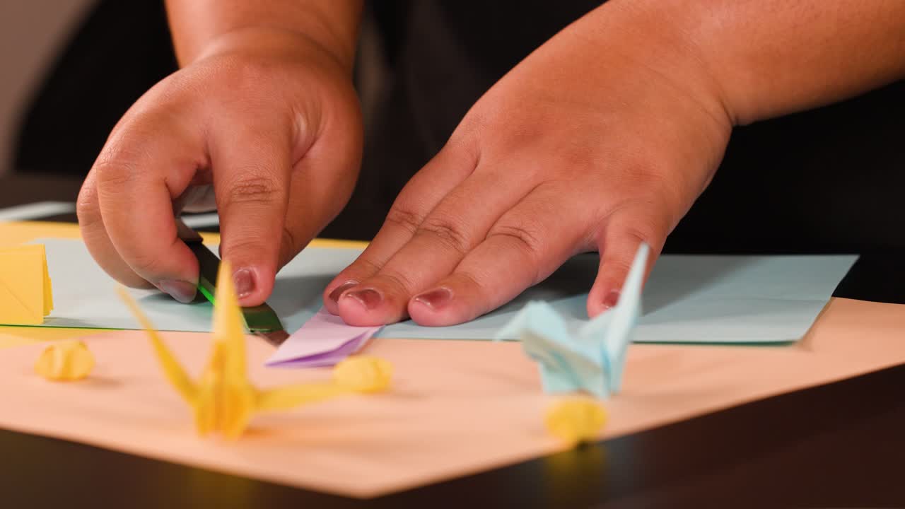 Close up shot of hands carefully folding pink paper on a desk with origami cranes