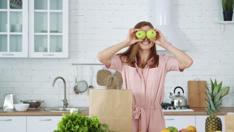 a smiling girl in the kitchen takes apples from a paper bag and puts it on her eyes