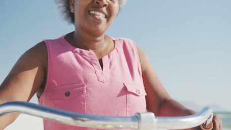 Happy-senior-african-american-woman-walking-with-bike-at-beach,-in-slow-motion