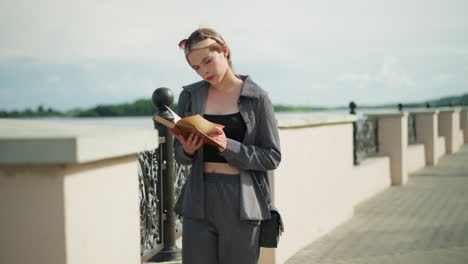 woman in grey clothing and black handbag stands by a fence, flipping to a new page in a book while focused, the wind blows through the pages with trees visible in the distance by the riverfront