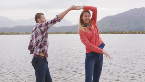 Caucasian-couple-having-a-good-time-on-a-trip-to-the-mountains,-dancing-near-lake-and-smiling