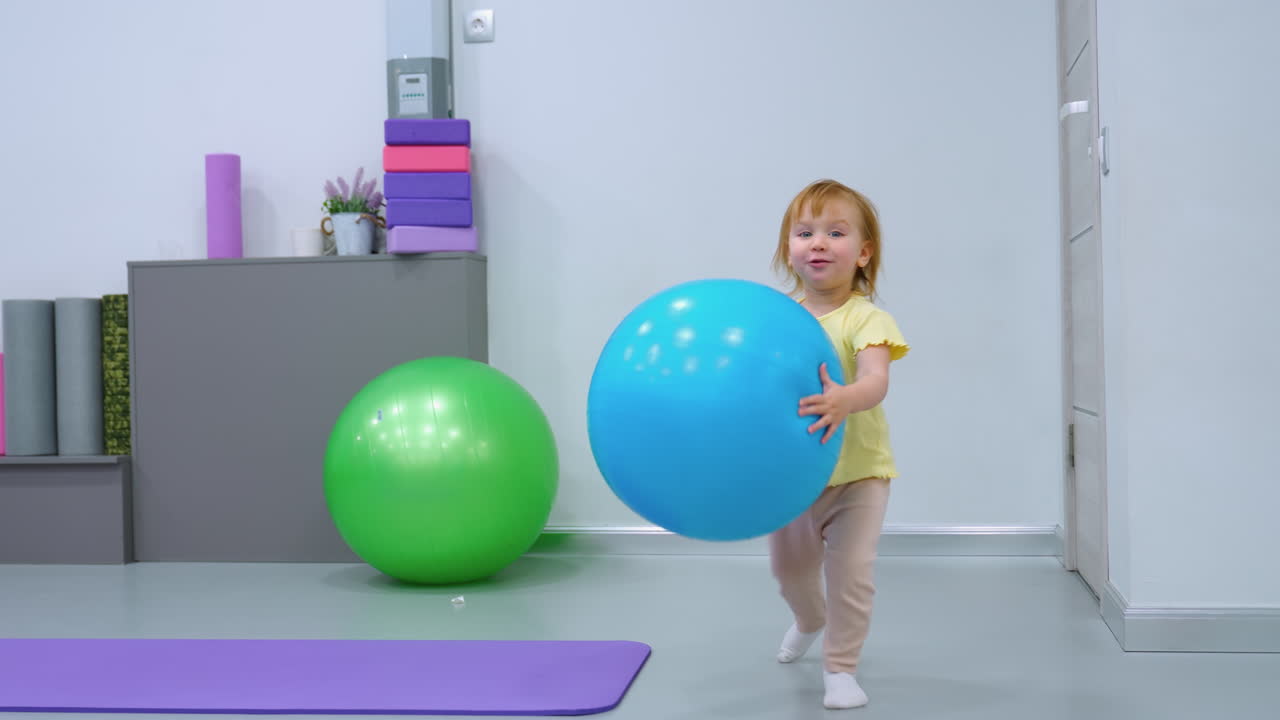 Seorang gadis kecil yang gembira mengulurkan tangan untuk meraih balon biru sambil berjalan tanpa alas kaki di lantai dalam ruangan, balon hijau berada di dekatnya, pemandangan penuh warna di ruang modern yang cerah dengan peralatan kebugaran.