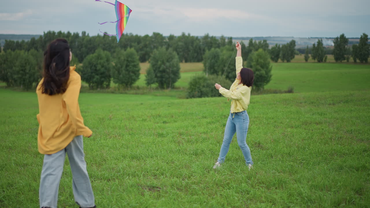 Dos mujeres vuelan una cometa colorida al aire libre con un cielo nublado sobre una pradera verde ondulada. Una lanza la cometa mientras la otra observa cerca, con ropa informal de mezclilla y chaqueta amarilla, movimiento lúdico en un viento suave.