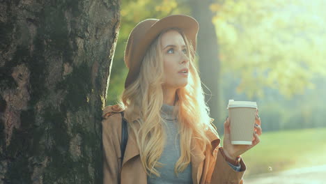 close-up view of blonde woman wearing hat, leaning on a tree, drinking a coffee to go, and looking around with a smile on her face in the park in autumn