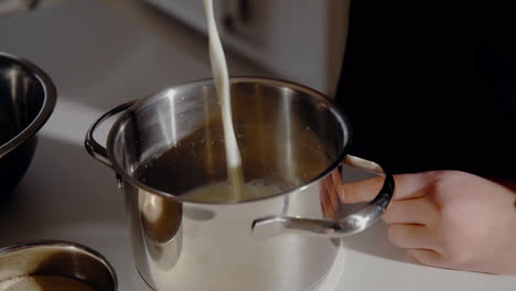 Mujer-Joven-Derramando-Leche-En-Una-Olla-Preparando-Masa-Para-Un-Pastel-En-La-Cocina-En-Casa