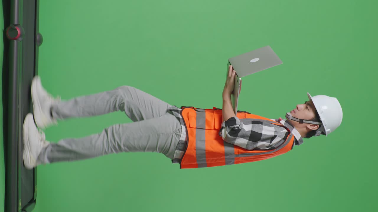 vista lateral de cuerpo completo ingeniero masculino asiático con casco de seguridad trabajando en una computadora portátil mientras camina en el estudio de fondo de pantalla verde