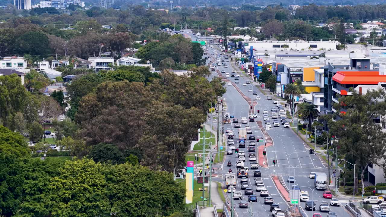 High-angle drone footage captures steady vehicle movement along a multi-lane city road bordered by trees and commercial buildings in daylight
