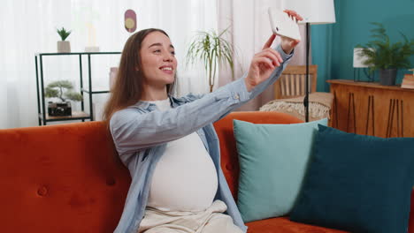 Mujer-Embarazada-Feliz-Sentada-En-El-Sofá-Tomándose-Un-Selfie-Con-Un-Teléfono-Inteligente-Tocando-Su-Barriga-Disfrutando-De-La-Maternidad