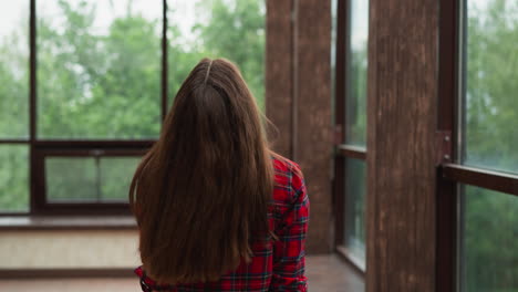 woman walks tilting head back along terrace. lady with long red hair wearing checkered shirt rests in studio with panoramic windows. luxury house