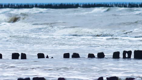 Stormy-waves-breaking-against-old-wooden-pier-on-the-beach,-overcast-spring-day,-Baltic-sea,-Latvia,-Pape,-medium-shot-from-a-distance