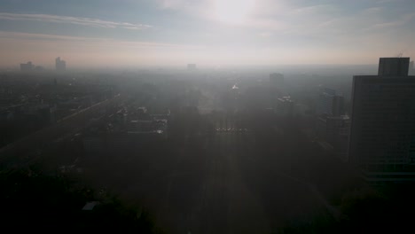 Drone-flight-against-the-sun-in-winter-Cologne,-Germany---Sun-rays-between-the-buildings-and-skyscrapers-in-the-background--flight-over-a-park