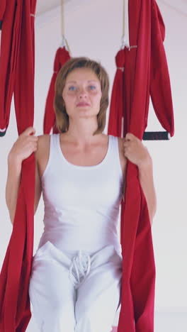 sporty lady wearing white sport suit relaxes after fly yoga workout sitting and swinging on red hammock close view