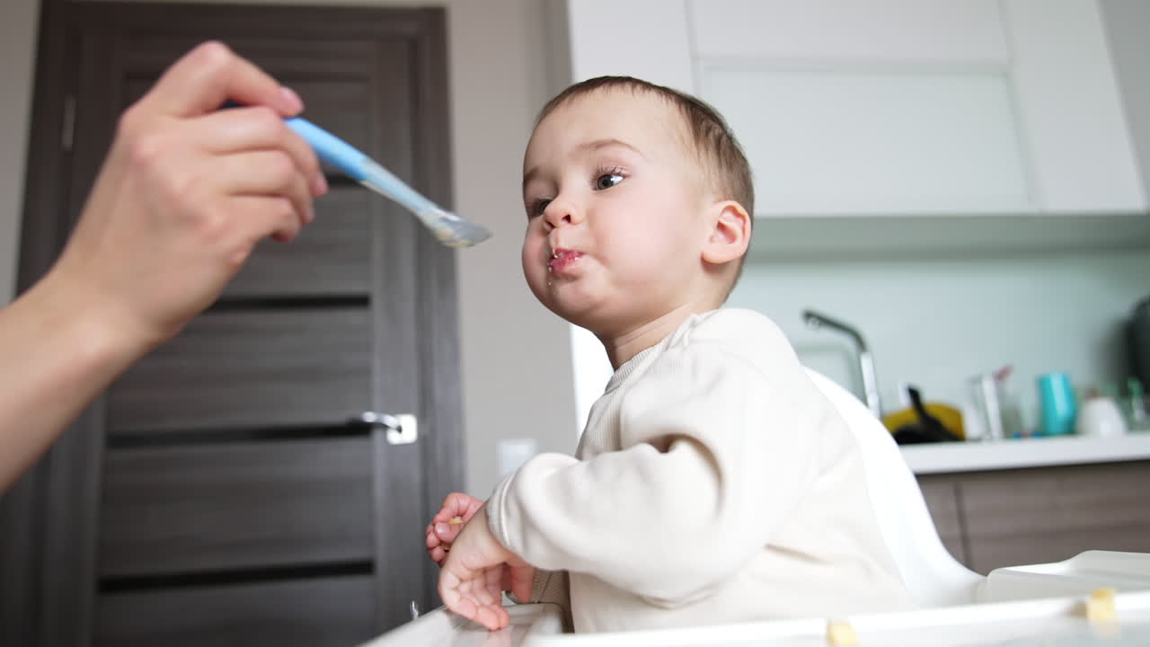 Sweet little toddler is fed from a spoon. Kid is chewing food slowly and takes a piece of cheese from table. Low angle view.