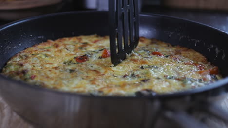 slow pan right to left of plastic spatchula cutting into cooked tomato, red pepper, kale, zucchini- courgette, bacon and onion omelette fritata in frying pan