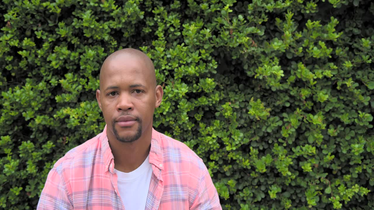Adult African man sitting by hedge shifting head torso returning gaze smiling pink plaid white tee