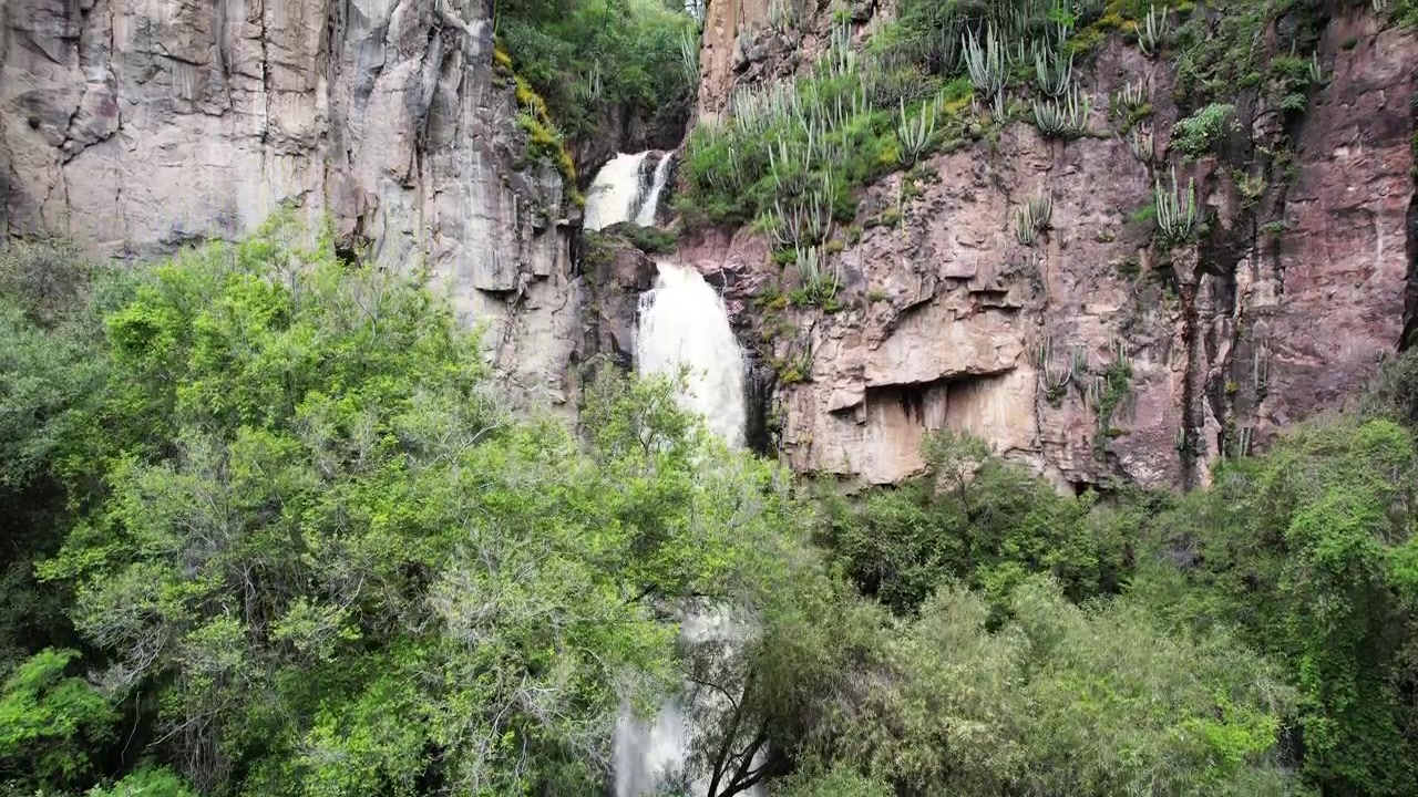Imágenes aéreas de dron que revelan una cascada fluyendo a través de acantilados rocosos y vegetación exuberante en Cerro Blanco, Aguascalientes, México. Impresionante paisaje natural rodeado de verdor y terreno accidentado.