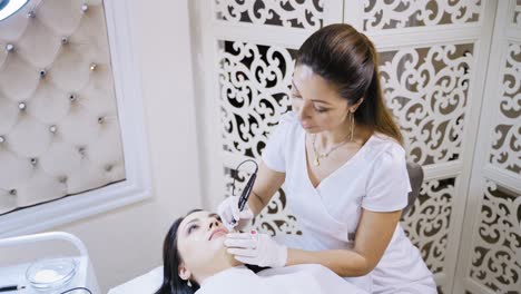 een elegante schoonheidsspecialiste in een labjas die met een cliënt praat tijdens een permanente lip make-upprocedure op de achtergrond van een witte decoratieve muur in een schoonheidssalon. close-up.