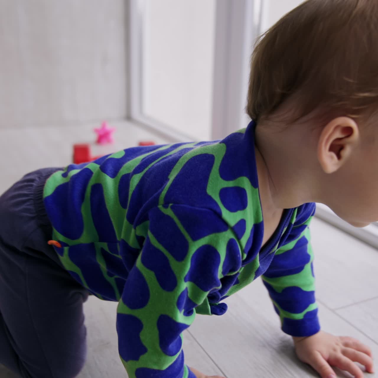 Nice cute toddler boy playing on the floor near the big windows. Kid wants to take off his sock but then crawls somewhere. Toys at backdrop in blur