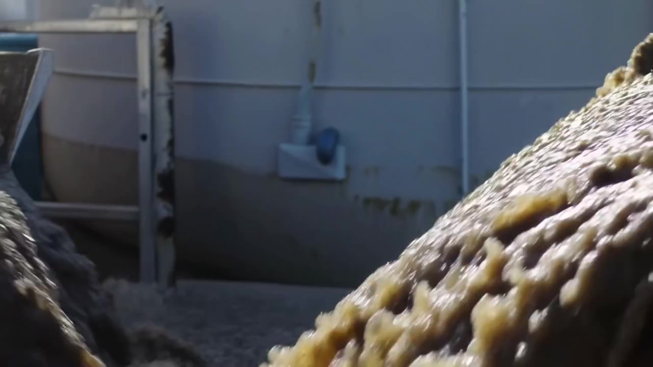 Close-up motion view of viscous brown slurry flowing over angled painted metal chute inside industrial processing facility, with glossy foamy texture, bright daylight reflections, wet surface, and