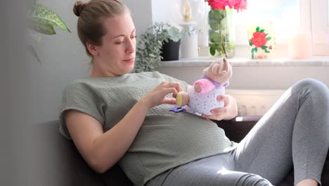 Pregnant-white-woman-sitting-on-the-couch-playing-with-a-baby-toy-at-sunlight-while-smiling,-slow-motion