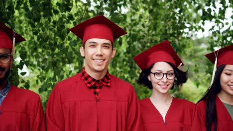 portrait of multinational group of graduating students in red graduation gowns and mortar-boards standing together outdoors, smiling and looking at camera.