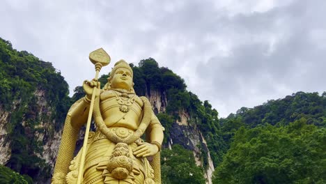 low angle shot of golden statue of hindu god murugan in front of subramanya temple with the view of steep mountain cliff in the background in malaysia