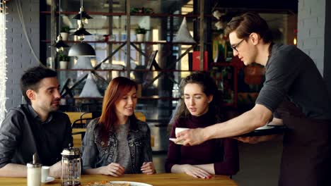 el amigable camarero está trayendo tazas de té a los estudiantes alegres sentados en la mesa en la cafetería. industria de alimentos y bebidas, comer fuera, conocer amigos y concepto de amistad.