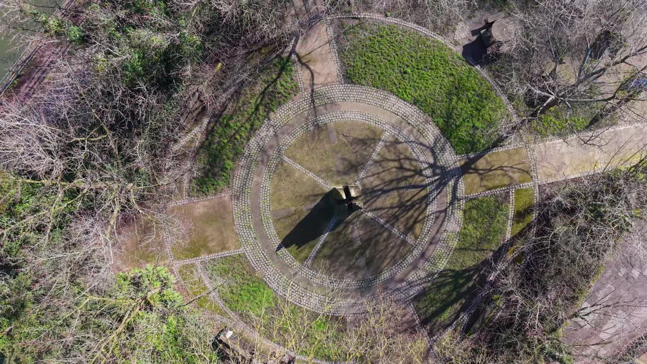 Stanstead Abbots monument aerial orbit with slight descent over circular paved plaza, winter trees and shadows in a quiet riverside village park, Hertfordshire, England, UK