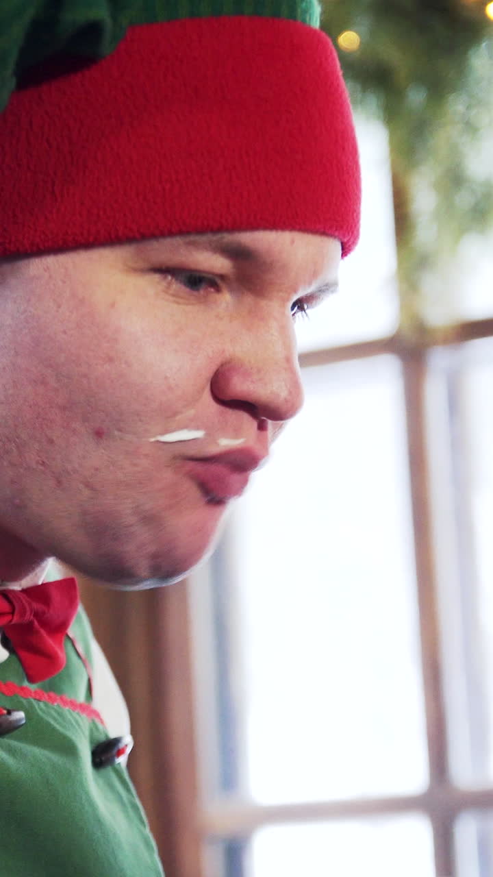 Funny elf with dirty face eats quickly. Hungry male elf in red and green hat eats and drinks indoors. Close-up. Vertical video