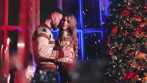 beautiful woman and her boyfriend indoors at christmas. lovely young couple in festive winter sweaters standing together near christmas tree at home.