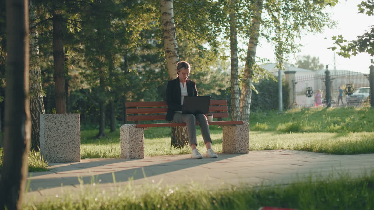 Remote worker seated outdoors on wooden bench using laptop and sipping coffee with lush green trees, sunlight, and passing bus in background