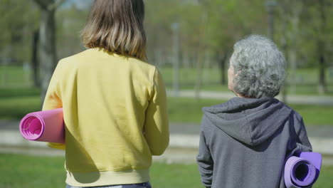 Vista-Trasera-De-Mujeres-En-El-Parque-Sosteniendo-Una-Estera-De-Yoga-En-Las-Manos,-Hablando