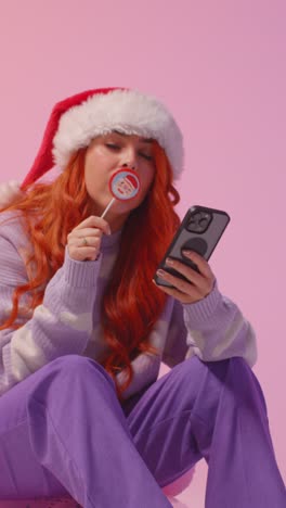 vídeo vertical de estúdio de jovem geração z mulher vestindo chapéu de papai noel comendo doces lollipop olhando para o telefone móvel