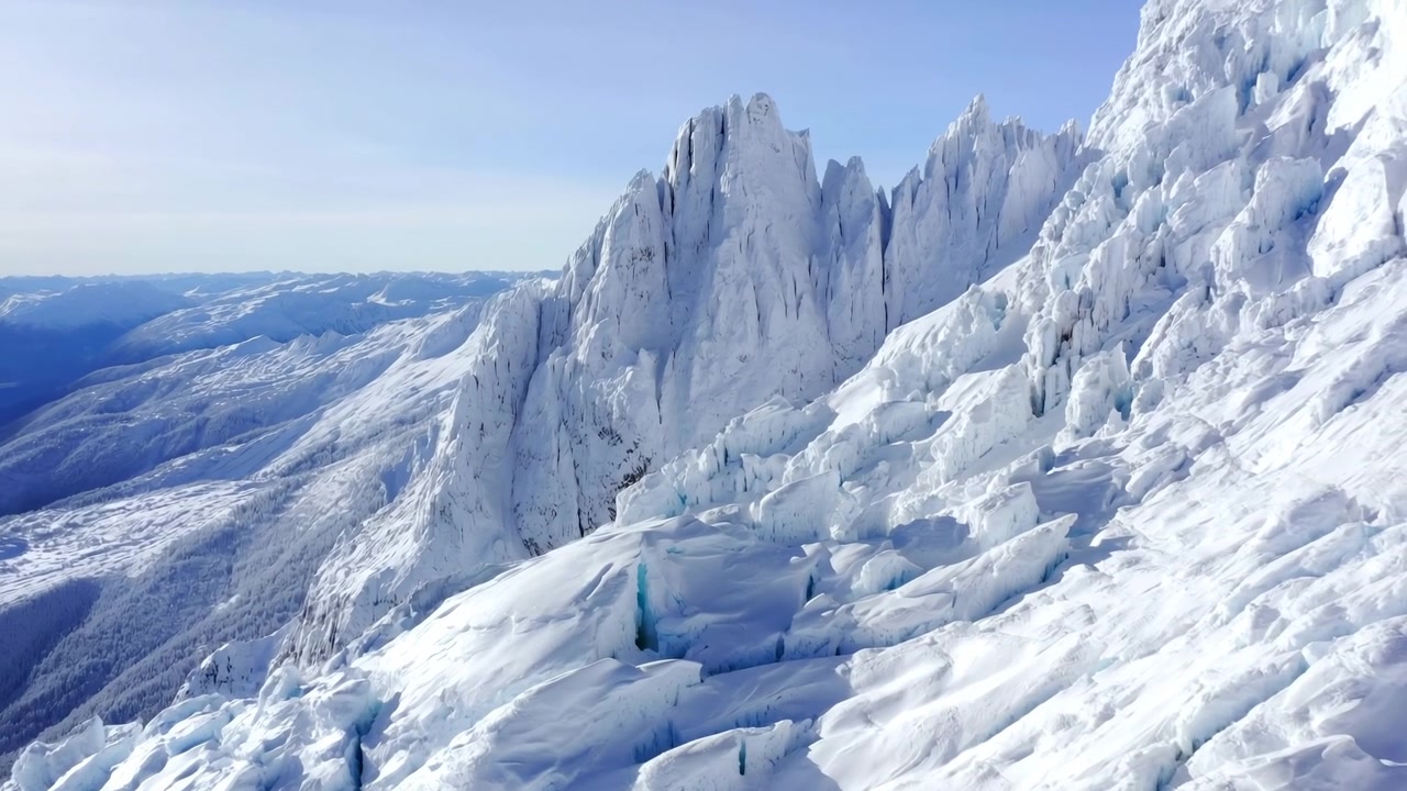 Aerial footage showing a sunlit high-altitude glacier with blue ice seracs, deep crevasses and steep snow-covered ridges under a clear blue sky