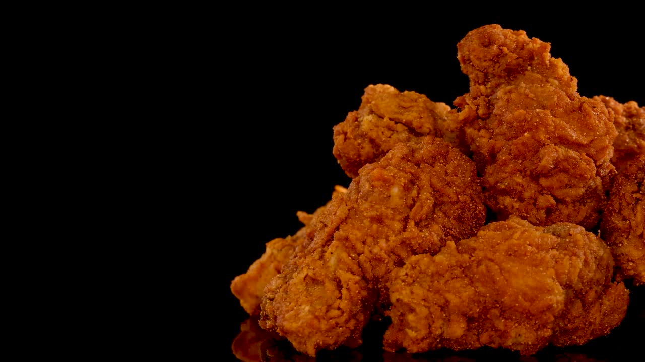 Crispy fried chicken pieces with spicy seasoning are slowly zoomed in on, under dramatic studio lighting, against a black background for a mouthwatering effect