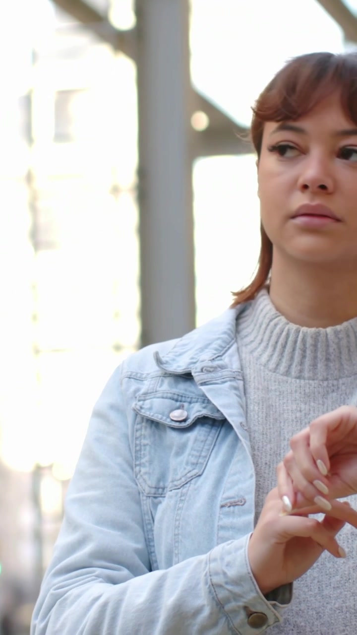 Video vertical: Mujer de pie con chaqueta vaquera revisando un anillo junto a una viga de metal en la calle, espacio para texto.