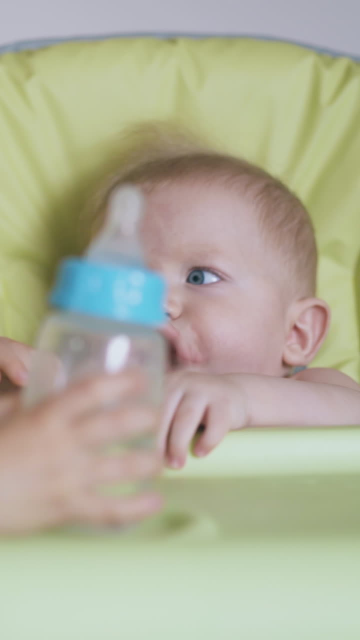 little blonde girl takes off bottle with water from junior brother sitting in green highchair in light room closeup